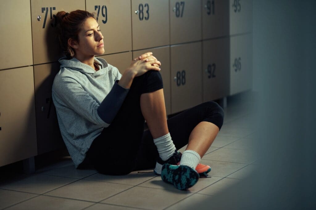 A distraught young woman sitting on the floor in a locker room, reflecting a deep sadness and contemplation, illustrating the emotional toll of sexual abuse in athletics.