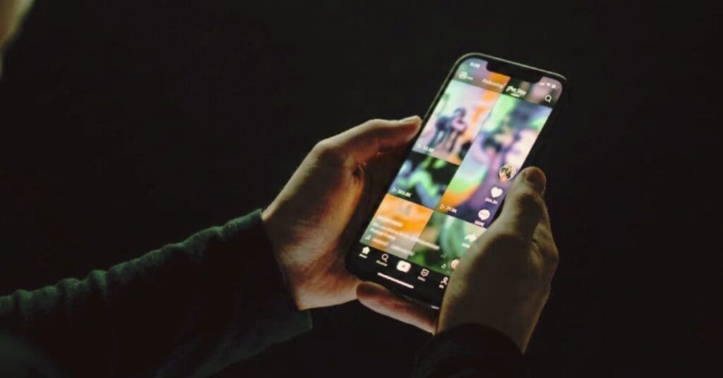 Hands holding a smartphone in a dark room showing a colorful, continuous vertical video interface, illustrating algorithmic compulsion.