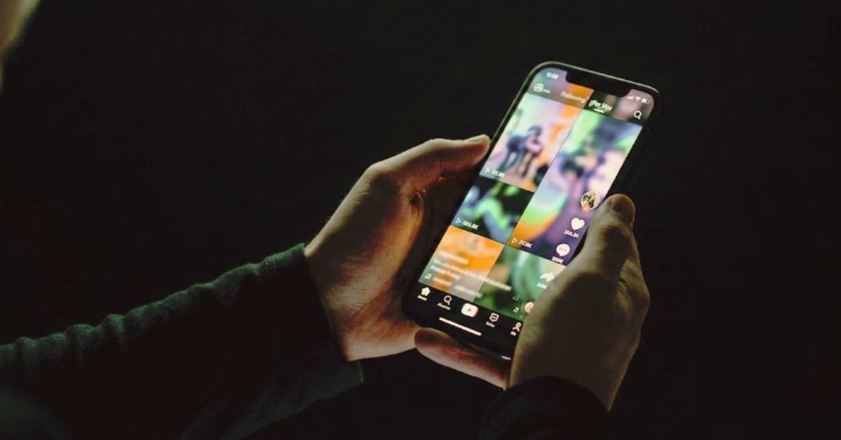 Hands holding a smartphone in a dark room showing a colorful, continuous vertical video interface, illustrating algorithmic compulsion.